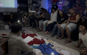 Fotografia de um encontro coletivo em ambiente escuro e artístico, com várias pessoas sentadas em círculo assistindo a uma apresentação projetada na parede. No centro da sala, tecidos coloridos, papéis e materiais estão espalhados no chão. Uma pessoa sentada em primeiro plano lê um texto enquanto o grupo observa atentamente. Ao fundo, há elementos visuais, cartazes e luzes neon que dão ao espaço um aspecto cultural e experimental.