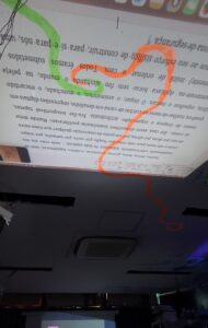 Fotografia de uma projeção em sala escura mostrando um texto em português exibido de cabeça para baixo no teto ou parede. Algumas palavras estão destacadas com marcações coloridas feitas digitalmente: uma linha verde circula a palavra “alinhando” e uma linha laranja serpenteia pela projeção. O ambiente tem iluminação baixa e teto com aparelho de ar-condicionado visível.